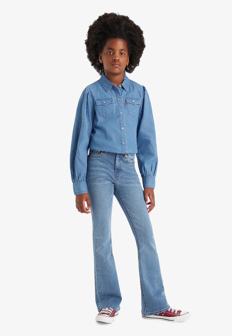 Blaue Denim-Hemdjacke mit Brusttaschen; kombiniert mit hellblauen Schlagjeans und roten Sneakern mit weißer Sohle.
