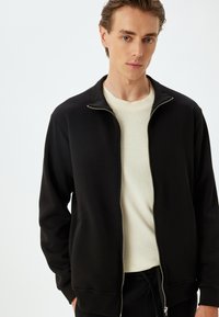 Svart zip-up jacka med ribbade krage och muddar, bärs över en krämfärgad tröja. Slät textur, minimalistisk design, figurnära siluett.