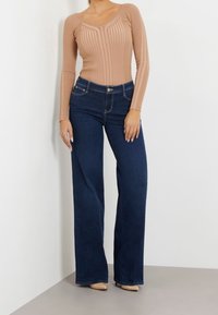 Braun geripptes Langarmoberteil mit V-Ausschnitt, kombiniert mit hochgeschnittenen dunkelblauen weit geschnittenen Jeans. Die Materialien wirken glatt und enganliegend.