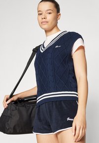 Gilet en maille torsadée bleu marine avec un col en V, avec des rayures blanches au col et à l'ourlet. Associé à un short bleu marine et à un sac noir.