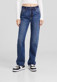 Weite blaue Jeans aus Denim mit hoher Taille, fünf Taschen und kontrastierenden Nähten. Getragen mit schwarz-weißen Sneakers.