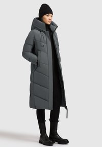 Lang grå pufferjakke med hætte, der har vandrette quiltede sektioner. Båret over sorte leggings og sorte ankelstøvler.