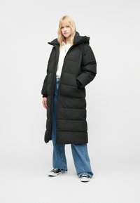 Fekete hosszú puffermellény cipzárral és kapucnival, steppelt mintával, oldalsó zsebekkel és laza fazonnal, kék széles lábú farmernadrág fölött viselve.