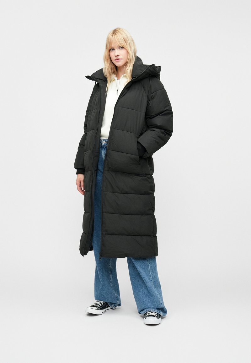 Fekete hosszú puffermellény cipzárral és kapucnival, steppelt mintával, oldalsó zsebekkel és laza fazonnal, kék széles lábú farmernadrág fölött viselve.