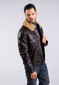 Braune Lederjacke mit einem hellbraunen Lammfellkragen, vorderen Taschen und Knopfverschlüssen. Kombiniert mit dunklen Jeans. Einfaches, klassisches Design.