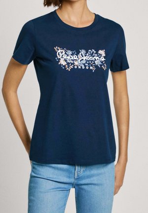 Kvinde iført en mørkeblå t-shirt med korte ærmer med blomsterprint "Pepe Jeans London" logo, kombineret med lyseblå denimjeans.
