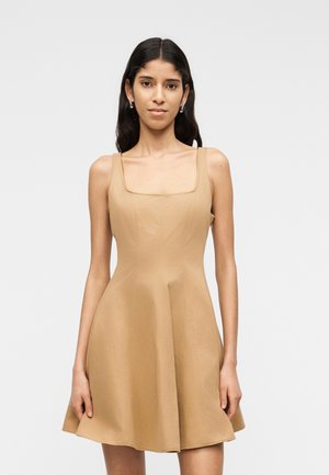 Kobieta o długich ciemnych włosach, ubrana w bezrękawnikową, beżową sukienkę z kwadratowym dekoltem, stojąca na tle jednolitego jasnego tła.