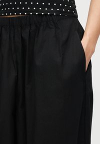 Κοντινό πλάνο ενός ατόμου που φορά μαύρο παντελόνι με ελαστική μέση και έχει το χέρι στην τσέπη, καθώς και μαύρο κοντομάνικο τοπ με λευκές πουά.