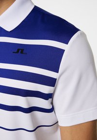 Pikétröja med horisontella blå och vita ränder, en svart logotyp och en klassisk krage. Tillverkad av ett mjukt, ventilerande tyg.