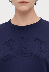 Sudadera azul marino con cuello redondo, que presenta un gran logo bordado "GAP". Tejido de algodón suave con una textura lisa.