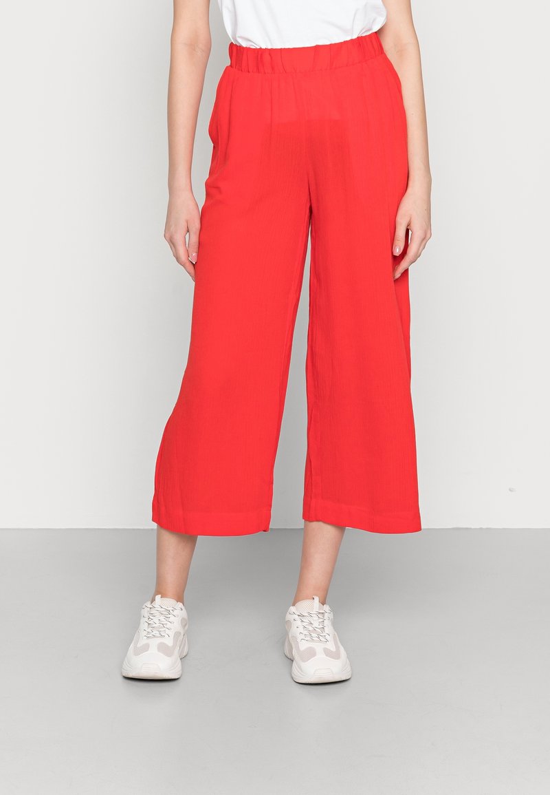 Femme portant un pantalon court large rouge vif avec une taille élastique et des baskets blanches, debout devant un fond uni.