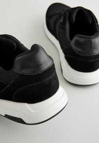 Schwarze Sportschuhe mit einem strukturierten Mesh-Obermaterial, Wildlederakzenten und einer weißen Zwischensohle. Die Ferse hat ein glattes Detail aus schwarzem Leder.