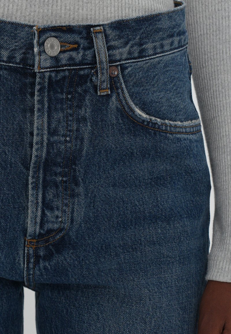 Blue denim jeans featuring a high waist, front pockets, and a metal button closure. Fabric shows a slight texture with prominent stitching details.
