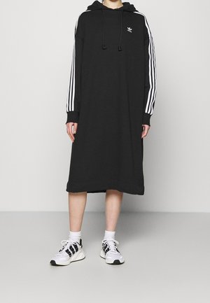Persona che indossa un vestito nero con cappuccio con strisce bianche lungo le maniche e scarpe da ginnastica bianche con dettagli neri, in piedi su un pavimento grigio.