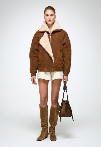 Bruine suède jas met een pluizige beige kraag, ritssluiting aan de voorkant en drukknopen. Model draagt beige shorts en bruine kniehoog laarzen, en houdt een tas vast.