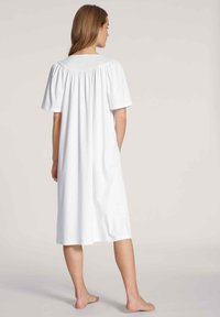 Un vestito da notte in cotone bianco con maniche corte, design fluido, dettaglio plissettato sul retro, lunghezza al ginocchio e tasche laterali. Collo piatto.