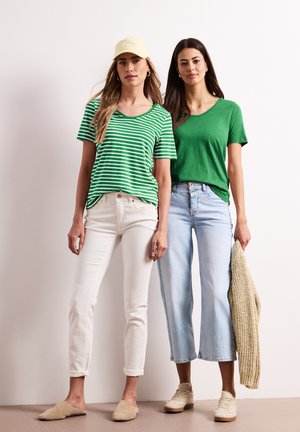 Dwie kobiety stoją obok siebie, ubrane w zielone topy; jedna w pasiastą koszulę, kremowe spodnie i beżowe mule z żółtą czapką, druga w jednokolorową zieloną koszulę, jasne dżinsy, trampki, trzyma sweter.