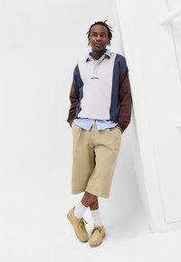 Πουλόβερ με μπλοκαρίσματα χρώματος σε γκρι, μπλε και καφέ, με γιακά και λεπτομέρεια κειμένου. Μπεζ σορτς και ανοιχτά παπούτσια, με λευκές κάλτσες.