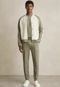 Chándal de color crema y verde oliva con botones a presión, combinado con pantalones elásticos a juego. Zapatillas blancas complementan el conjunto.