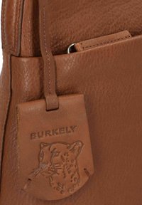 Angolo di una borsa in pelle marrone con un'etichetta del marchio Burkely, in rilievo, che presenta un design di testa di leopardop attaccata da una cinghia.