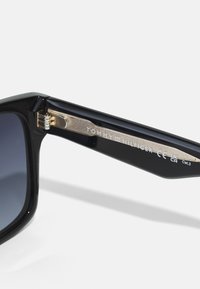 Svarte plast solbriller med mørke linser. Har metallarmer med inskripsjonen "TOMMY HILFIGER", og et hengsel for økt holdbarhet.