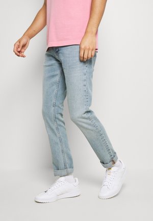 Personne portant un jean bleu clair retroussé, des baskets blanches, des chaussettes blanches et un t-shirt rose à manches courtes, debout devant un fond blanc.