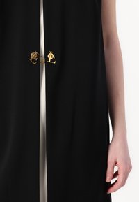 Roupa de tecido preto com uma abertura central, apresentando detalhes em metal dourado com letras entrelaçadas 'P' e 'L'. Textura suave e design minimalista.