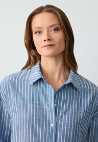 Light blue button-up shirt with vertical white stripes, made of a textured fabric, featuring a classic collar and light blue buttons.