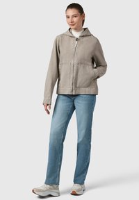 Giacca zip con cappuccio di colore grigio chiaro, due tasche frontali e cuciture a vista, abbinata a jeans blu e sneakers beige.