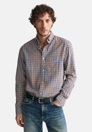 Camisa a cuadros en azul, marrón y blanco; con cuello abotonado; la tela tiene una textura suave; combinada con jeans oscuros y un cinturón negro.