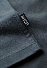 Mørkegråt stof med synlige syninger og et sort tøjlabel, der viser mærkenavnet "SUPERDRY" med hvide bogstaver.