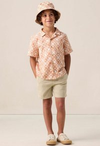 Korte mouwen overhemd met knopen in lichtoranje met witte patronen, gecombineerd met beige shorts en een bijpassende bucket hat; tan sneakers.