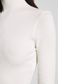 Haut à col roulé côtelé blanc, doté d'une coupe ajustée et d'un tissu texturé, mettant en valeur des lignes élancées et un col sans coutures.