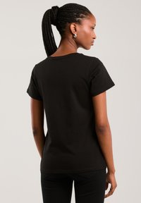 Femeie cu coadă de cal împletită, purtând un tricou simplu negru cu mâneci scurte și pantaloni negri, cu fața spre spate, pe un fundal neutru.