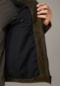 Indersiden af en brun ruskindjakke med sort stoffor og en lynlåslomme på venstre side, med en synlig metallynlås.