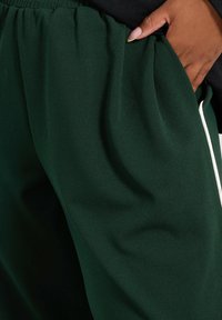 Gröna tygbyxor med sidorfickor, en slät textur och vita dragsnören vid midjan. Inga synliga mönster eller detaljer.