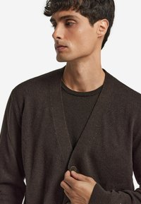 Mörkbrun cardigan med v-ringad design, mjuk textur och knapppdetaljer. Bärs över en matchande mörk skjorta.