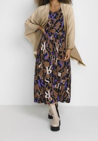 Brun midi-klänning med ett upprepat abstrakt bokstavsmönster i lila, svart och kräm. Bärs med en stickad beige sjal och beige knälånga stövlar.