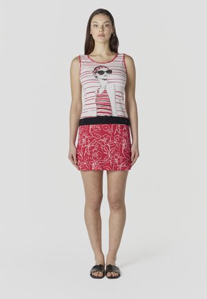 Femeie îmbrăcată într-un top fără mâneci cu dungi, având o ilustrație imprimată cu o femeie și o fustă roșie cu motive florale, stând în sandale negre pe un fundal alb.