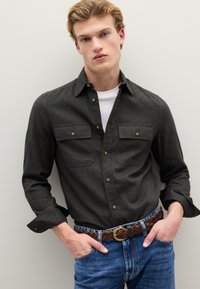 Donkergroen overhemd met knopen en twee borstzakken, gedragen over een wit T-shirt, gecombineerd met lichtblauwe spijkerbroek en een gevlochten bruine riem.