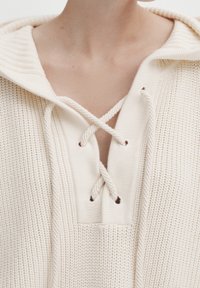 Crème gebreide hoodie met een gestructureerde stof, voorzien van een V-hals met kruisachtig vlechten en ribbelaccenten bij de kraag en de zoom.