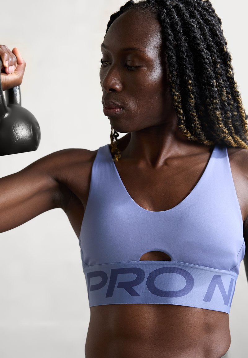 Mujer levantando una kettlebell negra con el brazo doblado, vistiendo un sujetador deportivo azul con texto, expresión concentrada, fondo claro.