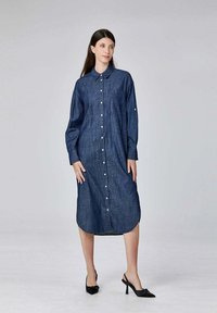 Femme portant une robe en denim à manches longues et boutons avec col, se tenant sur un fond uni, chaussée de talons décolletés noirs.