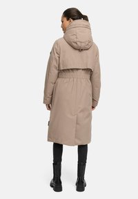 Manteau matelassé beige avec une partie supérieure courte et une ceinture élastique. Il dispose d'une grande capuche et de poches latérales, associé à un pantalon noir et des bottes.
