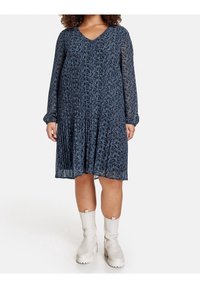 Abito blu navy con scollo a V, maniche lunghe trasparenti, design plissettato e motivo paisley. Indossato con stivali alla caviglia bianchi.