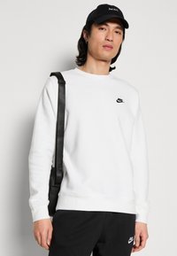Valkoinen collegepaita, jossa on pyöreä kaula-aukko ja ribbireunat, jossa on pieni musta Nike-logo. Yhdistetty mustiin housuihin ja mustaan lippikseen.