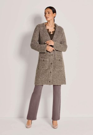 Femme portant un long cardigan en maille marron avec de grandes poches, un haut orné de dentelle, un pantalon gris et des talons beiges, debout contre un mur uni.