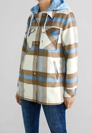 Persoon die een geruit jack draagt in bruin, blauw en wit met borstzakken en een lichtblauwe capuchon, gecombineerd met een blauwe spijkerbroek.