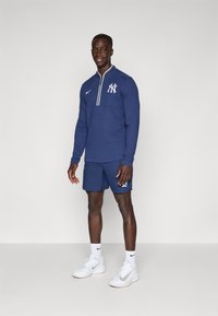 Námořně modrá košile s dlouhým rukávem na zip s bílým logem New York Yankees, spárovaná s námořně modrými šortkami, bílými ponožkami a bílými botami Nike.