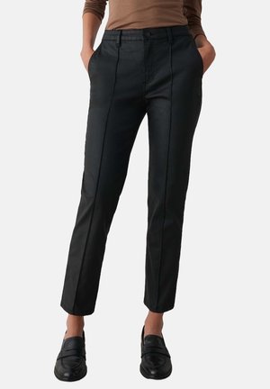 Kvinde iført sorte slim-fit bukser med forreste sømdetaljer, en brun langærmet top og sorte slip-on sko, hænderne i lommerne.